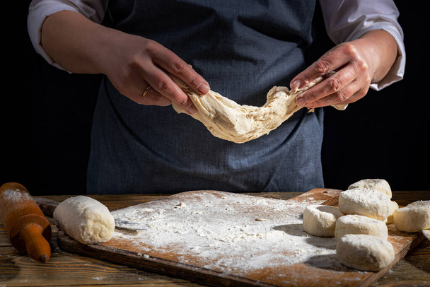 Female hands knead the dough on a wooden antique table on a dark background, close-up, shallow depth of field, beautiful directional lighting. Concept of home baking and comfort. - Foto, afbeelding