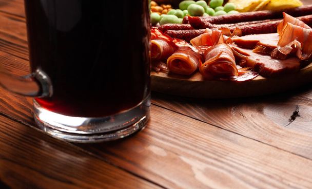 Mug of beer and assortment of snacks on a wooden table. Sausage, salami, ham, bacon. Beer salty snack, chips, wasabi nuts, salted peanuts, croutons. - Photo, Image