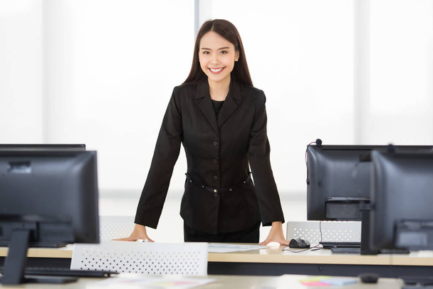コンピュータ画面と自信を持ってカメラに笑顔で現代のオフィスに立って正式な黒のスーツ姿の若いアジアの実業家を探してフレンドリーな。労働時間の幸せのためのアイデア. - 写真・画像