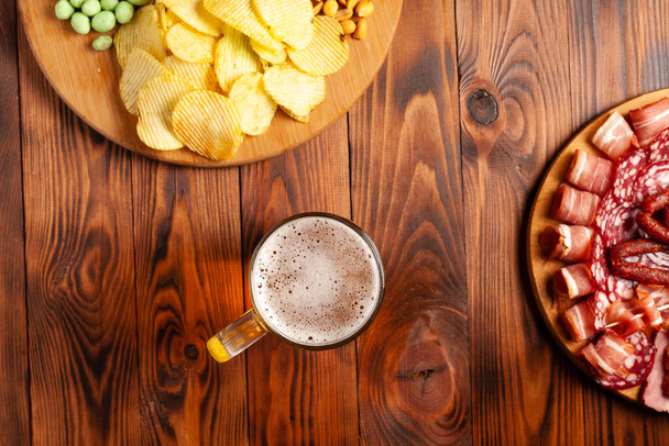 Mug of beer and assortment of snacks on a wooden table. Sausage, salami, ham, bacon. Beer salty snack, chips, wasabi nuts, salted peanuts, croutons. - 写真・画像