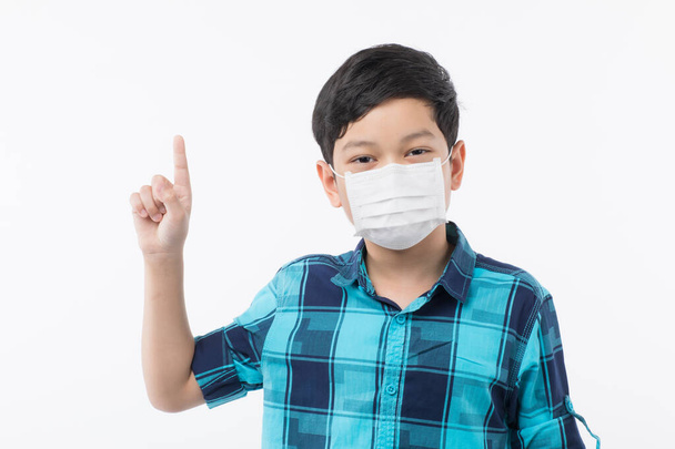 Jeune petit Asain garçon portant un masque de protection chirurgicale sur le visage sur fond blanc et regarder la caméra et le doigt pointeur vers spac noir
 - Photo, image