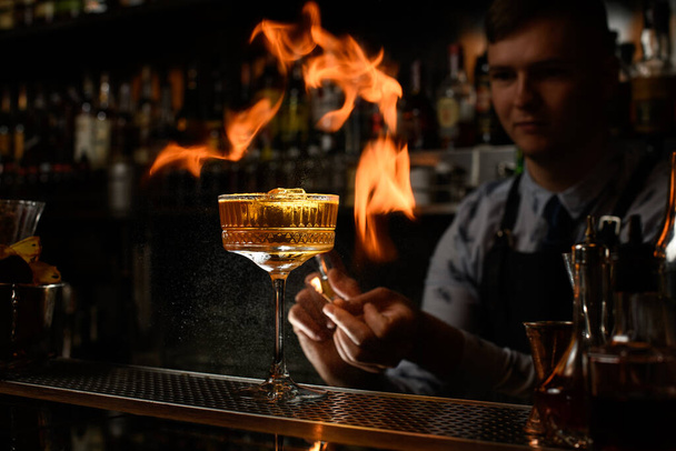 Junger Barkeeper sprüht sorgfältig und macht Feuer über Glas mit Cocktail - Foto, Bild