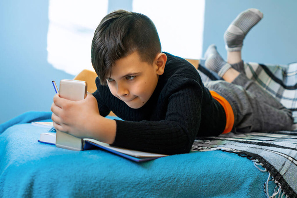 Distance learning online education. A schoolboy studies at home from the phone and does the homework of the school. Training books and notebooks on the table - Fotografie, Obrázek