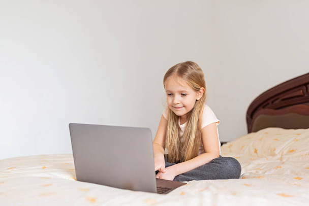 Stay at home quarantine coronavirus pandemic prevention. Little girl with blonde hair sitting on the bed and using laptop. Prevention epidemic. Child using technology - Photo, image