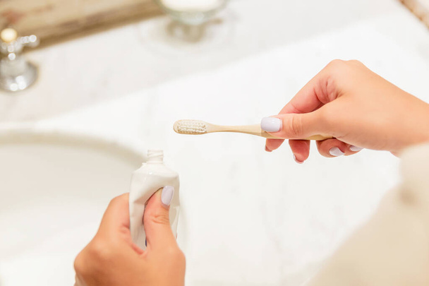 Imagen recortada de manos femeninas poniendo pasta de dientes en el cepillo de dientes en el baño. Concepto de cuidado dental. Preparación para la limpieza de dientes
. - Foto, imagen