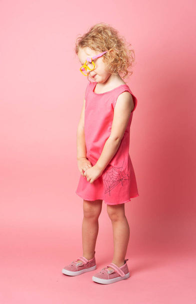 Little curly girl in funny glasses shows a pink dress. Portrait on a white background. - Foto, Imagen