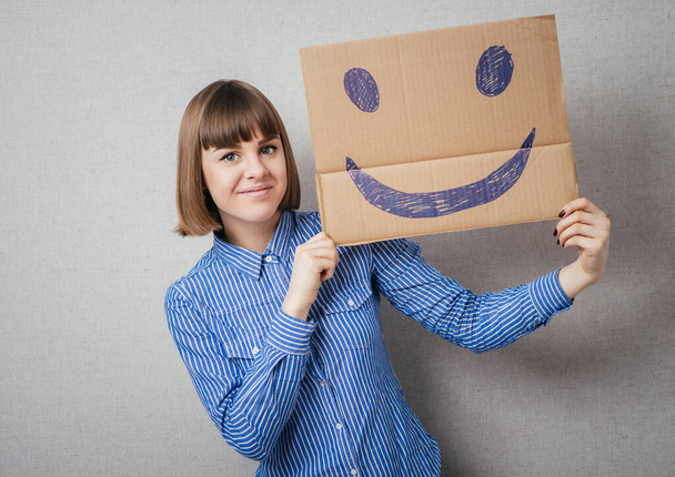 Giovane donna che tiene un cartone con una faccina sorridente davanti alla testa
. - Foto, immagini