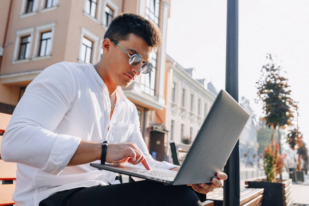 молодой стильный парень в белой рубашке с телефоном и ноутбуком работает на скамейке в солнечный теплый день на открытом воздухе, фрилансер
 - Фото, изображение