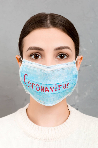 Portret zdenerwowanej kobiety w masce medycznej z napisem koronawirusowym na szarym tle cementu. Koncepcja koronawirusa. Ochrona dróg oddechowych. - Zdjęcie, obraz