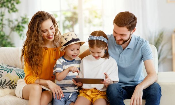 Bambina che condivide tablet con fratello e genitori sorridenti mentre si riuniscono tutti insieme sul divano in accogliente salotto
 - Foto, immagini