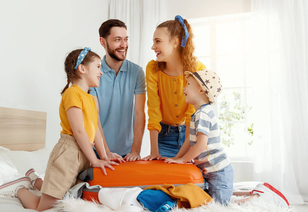 Riendo niños y padres reuniéndose en la cama cerrando la maleta juntos mientras se preparan para las vacaciones mirándose con sonrisa
 - Foto, Imagen