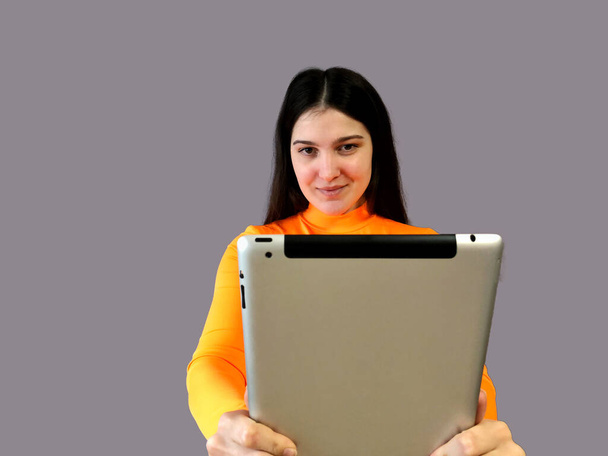 Young beautiful girl with dark hair in a bright orange sweater on a gray background. With electronic tablet in hands - Valokuva, kuva