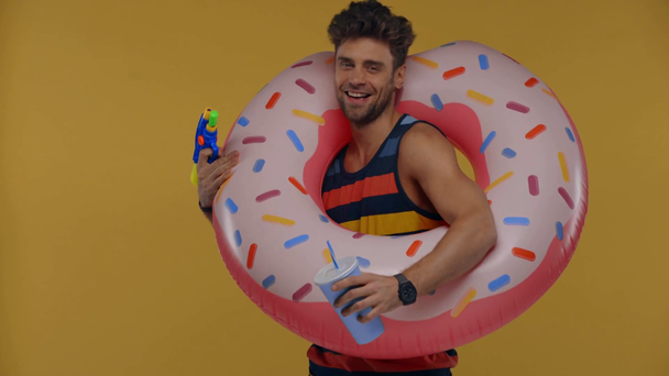 Homem sorridente com anel de natação, copo de papel e arma de brinquedo isolado no amarelo
 - Filmagem, Vídeo