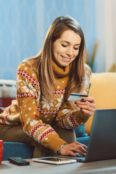 Happy young woman sitting on the couch at home and doing online shopping for Christmas gifts, she is connecting with her laptop and holding a credit card - Φωτογραφία, εικόνα
