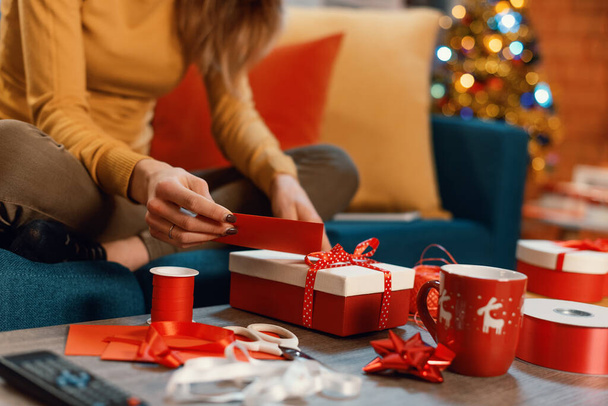 Woman preparing Christmas gifts at home: she is sitting on the sofa and adding a Christmas card to a present - Фото, зображення