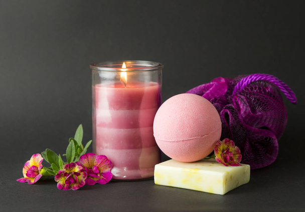 Wellness spa therapy pampering products on black background, pink candle burning, warm mood. Pink bath bomb, hand made soap, sponge, pink burning candle, flowers for decoration. Spa wellness concept. - Photo, Image