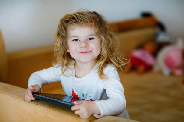 Симпатичная маленькая девочка, играющая дома с планшетным компьютером. Здоровый ребенок трогает тачпад пальцами, смотрит мультики и развлекается развивающими играми на компьютере. на карантине против коронавируса
 - Фото, изображение