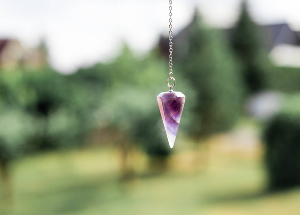 Purple natural Amethyst crystal quartz pendulum hanging on chain on green outdoors nature background. - Photo, Image
