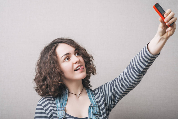 Woman playfully makes selfie on the phone, a grimace. Gray background - Fotografie, Obrázek
