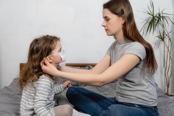 Moeder helpt haar dochter gekleed virus masker zittend op bed thuis, proberen om familie te beschermen tegen besmettelijke ziekten. Concept van coronavirus of COVID-19 pandemische ziekteverschijnselen, virale infecties - Foto, afbeelding