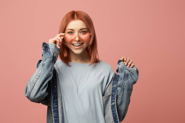 Happy woman in pink glasses and denim jacket standing in studio - Photo, image