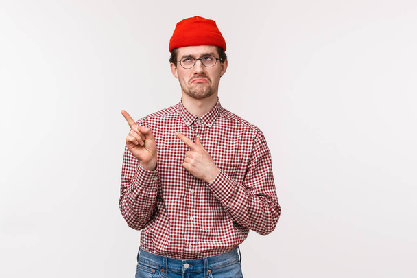 Sceptyczny i niezabawny, znudzony młody hipster w czerwonej czapce i okularach, zirytowany wizytą na nudnym spotkaniu, wskazywaniem lewego górnego rogu, przewracaniem oczami i dąsaniem się - Zdjęcie, obraz