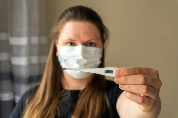 Une fille dans un masque médical tient un thermomètre dans sa main, qui montre une valeur élevée de la température corporelle. Symptômes du coronavirus. Travail à distance à domicile pendant une pandémie
. - Photo, image