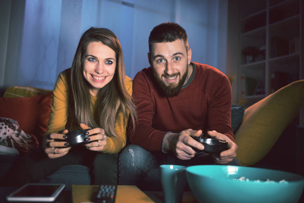 Happy couple playing video games together at home, they are sitting on the sofa and holding a game controller, entertainment and lifestyle concept - Foto, Bild