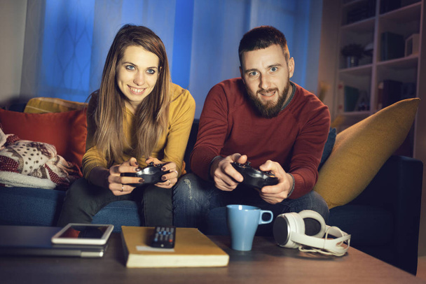 Happy couple playing video games together at home, they are sitting on the sofa and holding a game controller, entertainment and lifestyle concept - Fotografie, Obrázek