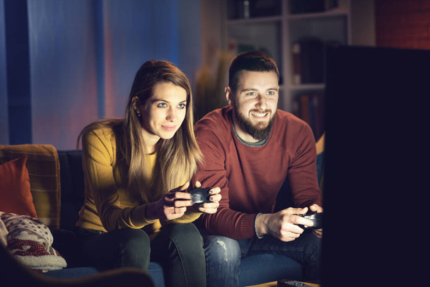 Happy couple playing video games together at home, they are sitting on the sofa and holding a game controller, entertainment and lifestyle concept - Фото, изображение
