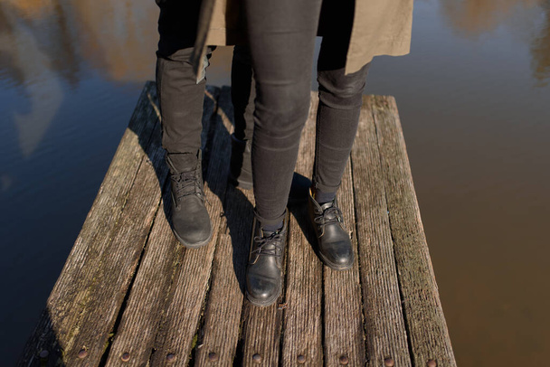 スニーカーで作物の写真の若い足、湖の近くの木製の橋の上に夫婦、夫と妻と結婚した。桟橋に立っているカップルの背面図。下半身。テキストとデザインのための場所。恋の夏. - 写真・画像