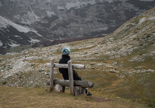 Молода жінка з блакитним волоссям у чорному пальто сидить на дерев'яній лавці на узбережжі гори
 - Фото, зображення