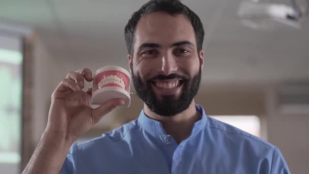 Close-up portrait of young Middle Eastern man holding teeth cast and smiling at camera. Professional orthodontist posing in dental clinic. Dentistry, medicine, employment, health care. - Video, Çekim