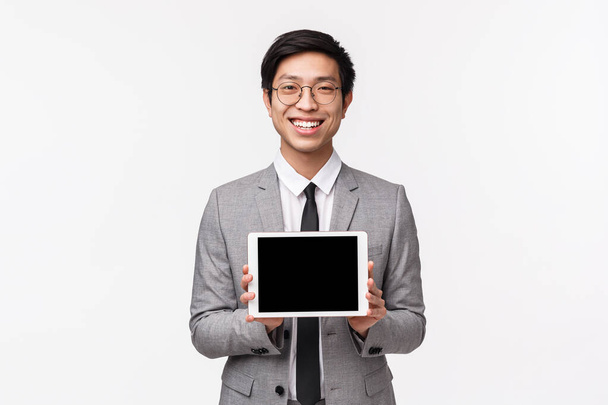 Taille-up portret van tevreden, goed uitziende Aziatische man in grijs pak en bril, introduceren toepassing, project of financiële presnetatie, tonen digitale tablet scherm en glimlachen tevreden - Foto, afbeelding