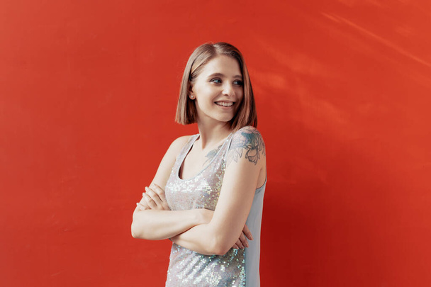 Veselá mladá dívka pózná před červenou zdí. Přirozené světlo. Tetování na levém rameni. Oblečené v lesklých šatech - Fotografie, Obrázek