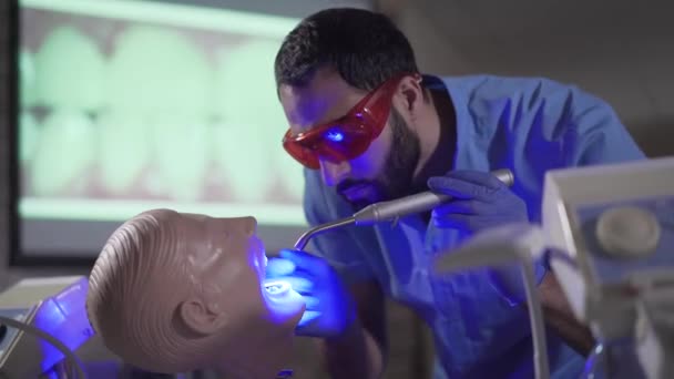 Portrait of busy Middle Eastern stomatologist assistant practicing with dental mannequin. Focused young man in protective eyeglasses using curing lights. Profession, medicine, stomatology. - Filmagem, Vídeo