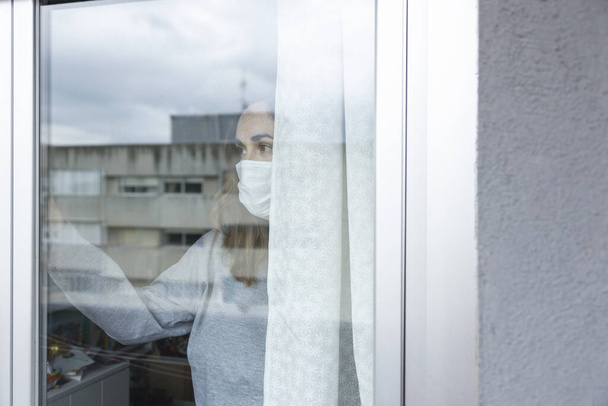 Nainen, jolla on hengityssuojain ja älypuhelin, katselee talonsa ikkunasta epävarmana, odottaen karanteenin päättyvän covid-19: llä
 - Valokuva, kuva