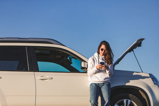 vrouw in de buurt van auto met geopende kap op zonsondergang praten op telefoon kopieerruimte - Foto, afbeelding