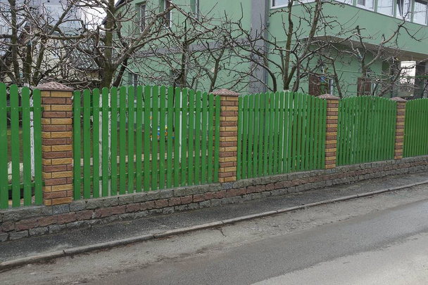 Gri asfalt bir yolun kenarında, uzun yeşil tahta ve kahverengi tuğlalardan oluşan özel bir çit. - Fotoğraf, Görsel