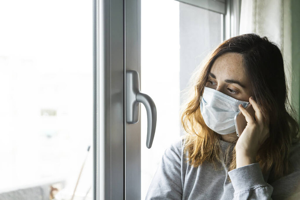 woman with respirator talking with her smart phone looking through the window of her house with uncertainty, waiting for the quarantine to end by covid-19 - 写真・画像