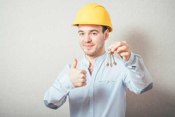 The man in yellow helmet holding keys and showing thumb up. On a gray background. - Photo, Image