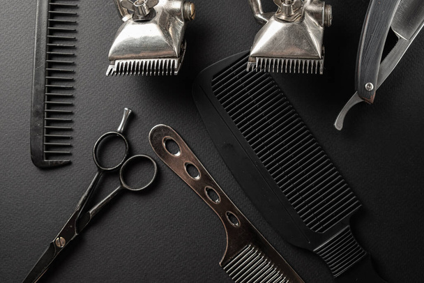 Auf einer schwarzen Oberfläche befinden sich alte Friseurwerkzeuge. Zwei alte Haarschneidemaschinen, Kämme, Rasiermesser, Friseurschere, Rasierpinsel. schwarz monochrom. horizontale Orientierung. Draufsicht, flache Lage - Foto, Bild