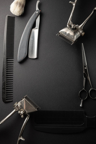 Auf einer schwarzen Oberfläche befinden sich alte Friseurwerkzeuge. Zwei alte Haarschneidemaschinen, Kämme, Rasiermesser, Friseurschere, Rasierpinsel. schwarz monochrom. vertikale Orientierung. Draufsicht, flache Lage - Foto, Bild