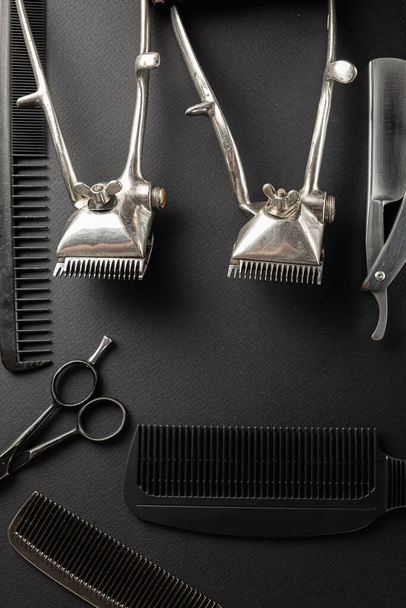 Auf einer schwarzen Oberfläche befinden sich alte Friseurwerkzeuge. Zwei alte Haarschneidemaschinen, Kämme, Rasiermesser, Friseurschere, Rasierpinsel. schwarz monochrom. vertikale Orientierung. Draufsicht, flache Lage - Foto, Bild