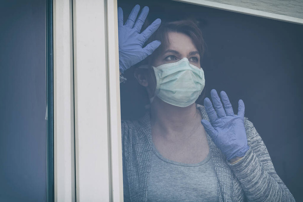 Woman wearing a protective face mask and looking out the window of her home because of an epidemic of corona virus covid-19. Home quarantine and self isolation concept - Photo, image