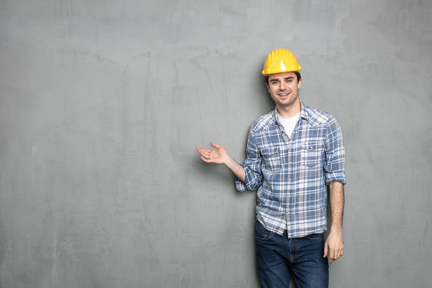 confident young worker with safety helmet presenting one empty side of the image for text or promoting something, on a concrete grey wall - Φωτογραφία, εικόνα