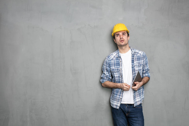 подумайте, прежде чем действовать, молодой работник со шлемом безопасности и современный планшет ищет решение, чтобы закончить проект, который он имеет, стоя на бетонной стене
 - Фото, изображение
