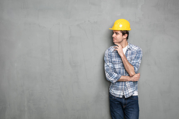 confident young worker with safety helmet presenting one empty side of the image for text or promoting something, on a concrete grey wall - Zdjęcie, obraz