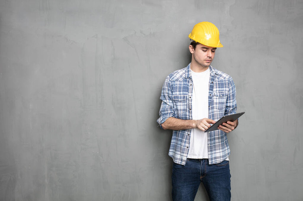 mladý inženýr pracující a dívající se na tablet pro projekt, který potřebuje dokončit, stojící na průmyslové betonové zdi - Fotografie, Obrázek