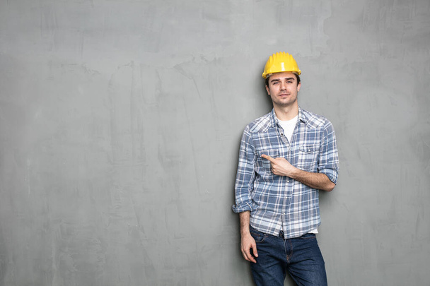 confident young worker with safety helmet presenting one empty side of the image for text or promoting something, on a concrete grey wall - Photo, image